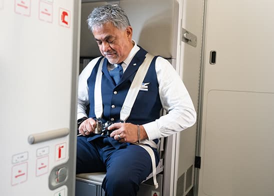 Grey-haired male flight attendant sitting down and putting on his seatbelt.